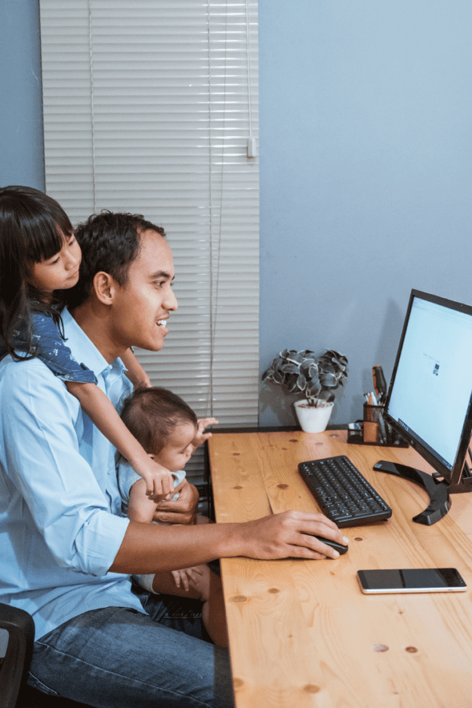 Asian man working from home while holding a baby and daughter hugging him from nehind