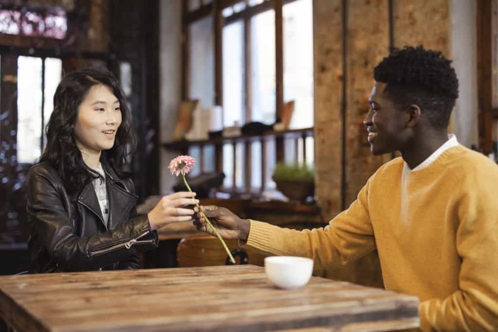 Black man wearing a yellow sweatshirt handing over a stem daisy to an Asian woman wearing black leather jacket on a date