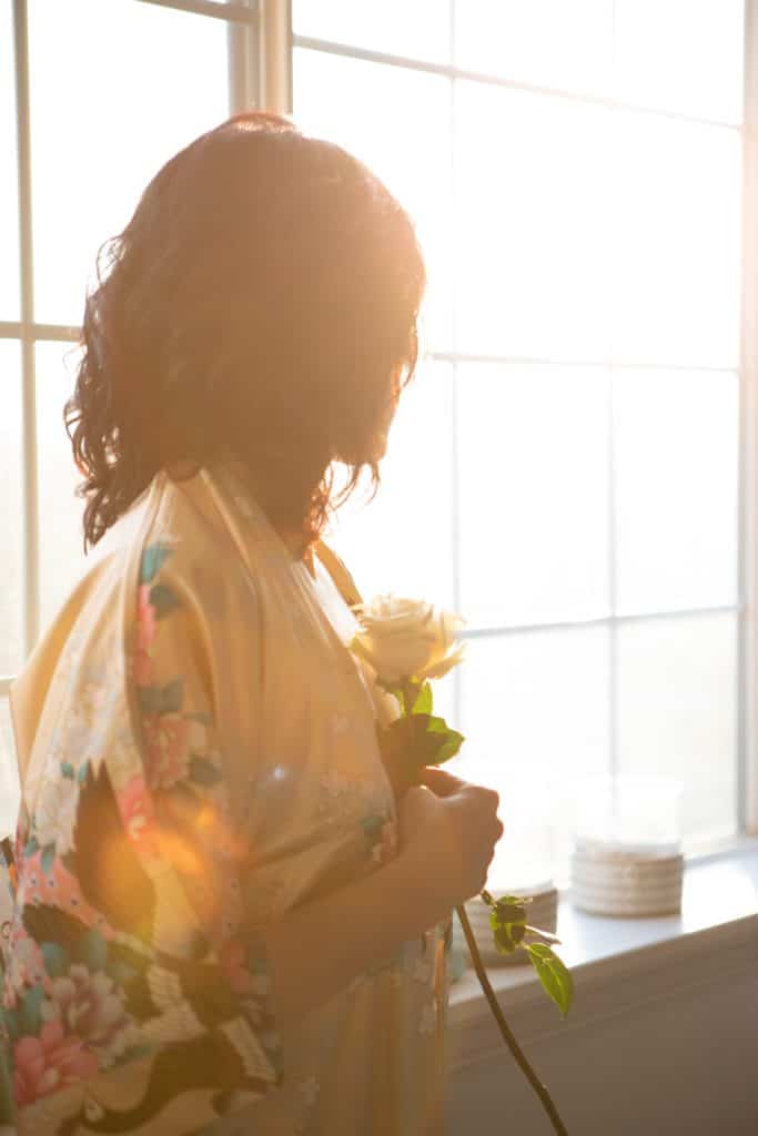 woman wearing a satin with printed flowers bathrobe holding a white stem rose looking outside the window