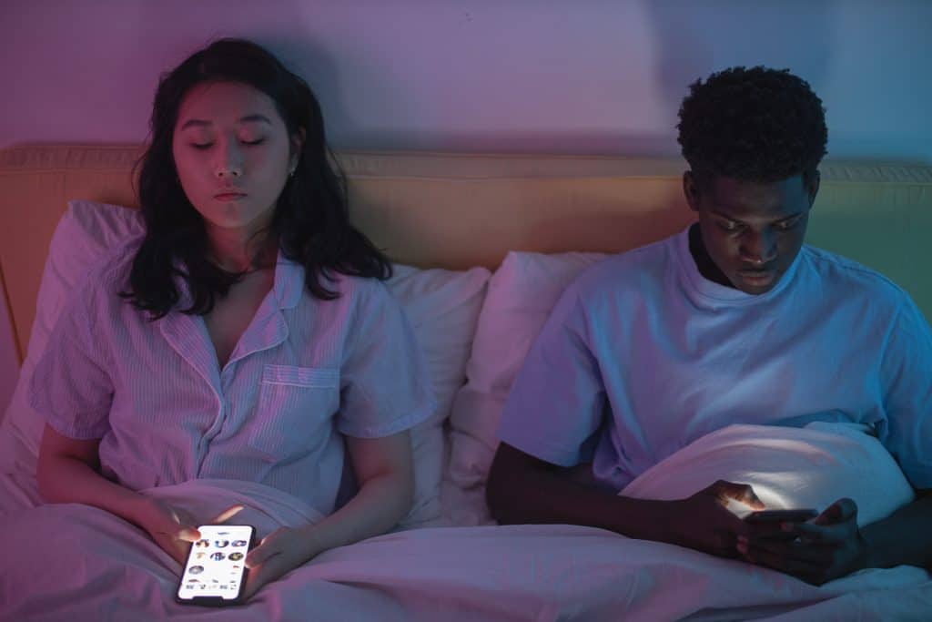 a multi-racial couple sitting on the bed holding their own phones