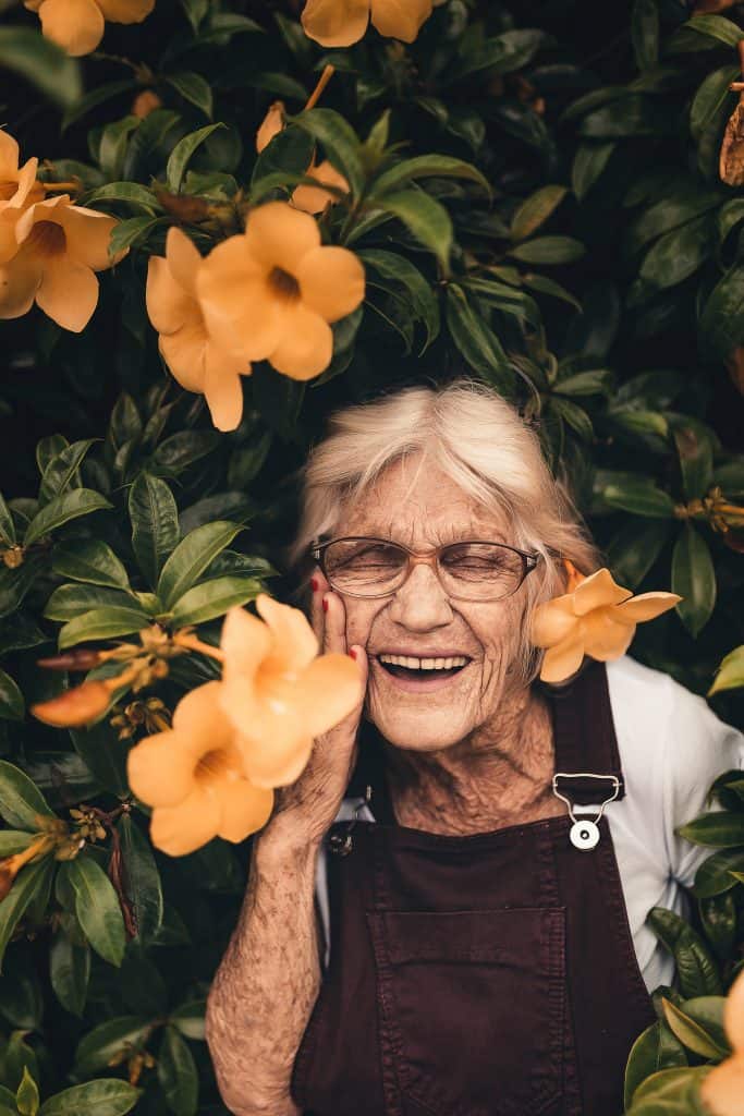 a happy old grey-haired woman wearing eye glasses in a bush of flowers