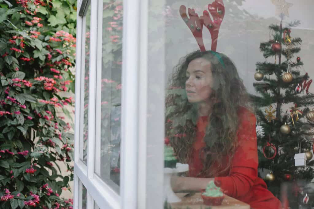 a photo of a pensive woman wearing a reindeer headband and red clothes looking outside the window during holidays and a Christmas tree behind her